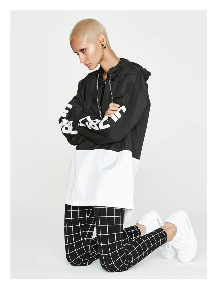 A woman with a shaved head and earrings is kneeling on the floor, wearing a black and white color-block windbreaker, black pants with white grid pattern, and white sneakers