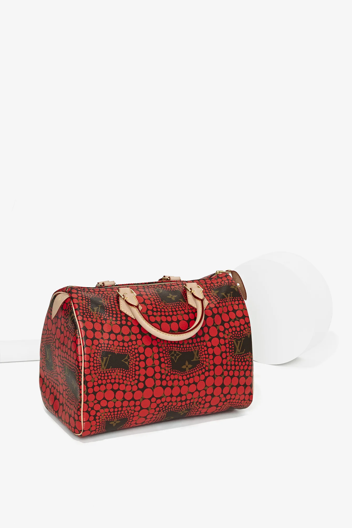 Red designer handbag with a pattern of circles and Louis Vuitton logo, placed on a white surface beside a white spherical object.