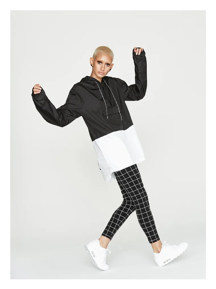 Person with short platinum blonde hair wearing a black and white windbreaker, black and white checkered pants, and white sneakers, posing against a plain white background.