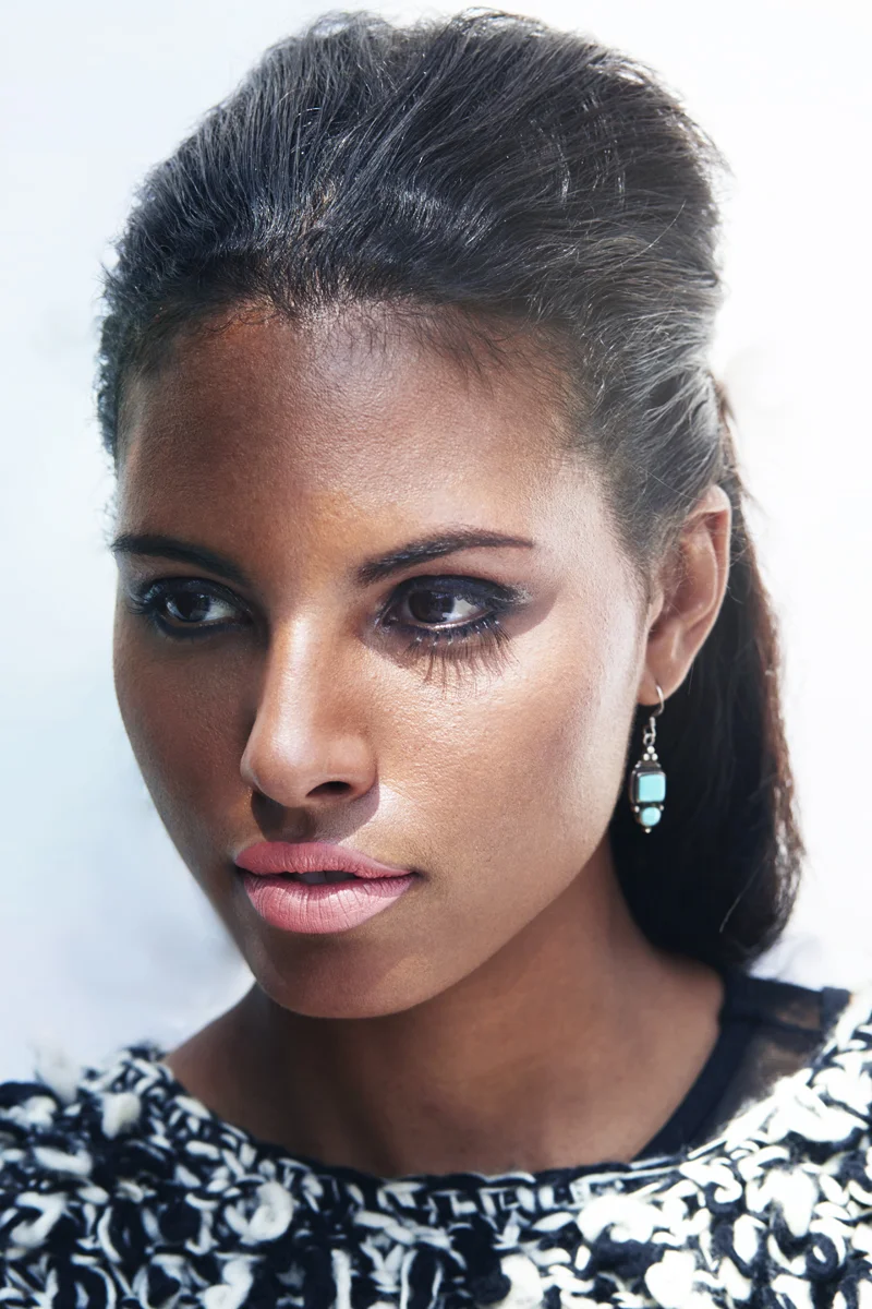 A woman with dark hair styled back, wearing earrings and a black and white patterned top, looking off to the side