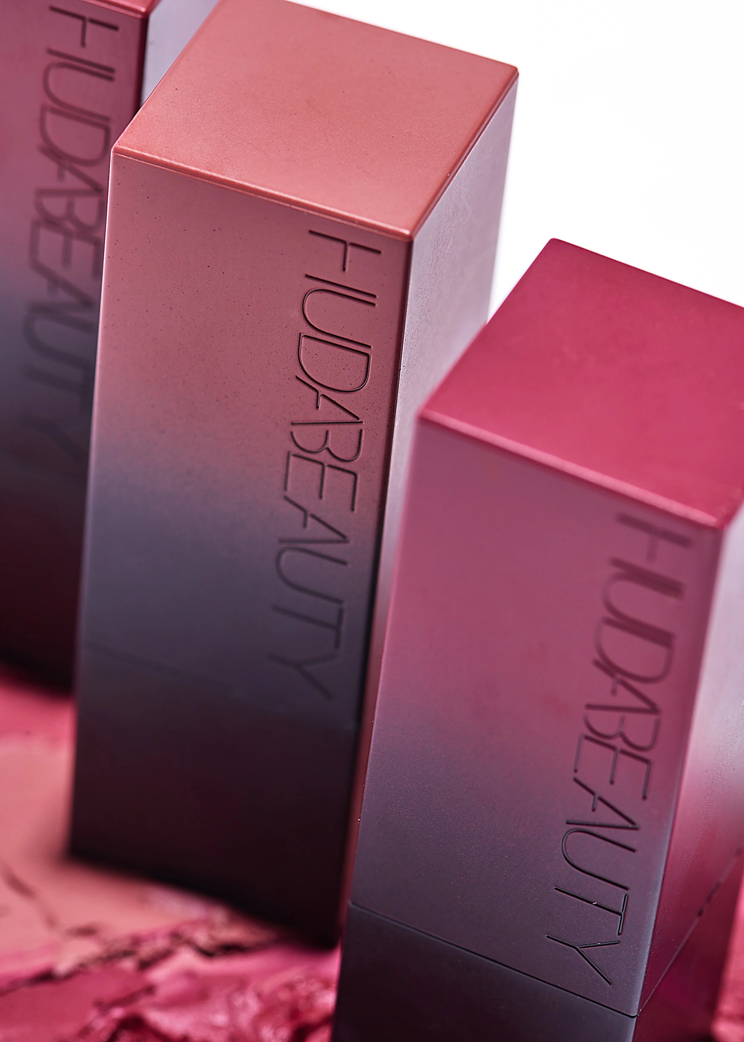 Close-up of three rectangular lipstick boxes with pink and metallic finishes, embossed with the words 'Huda Beauty'.