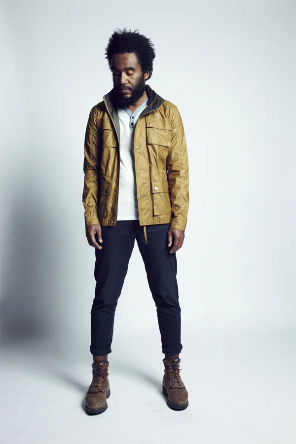 Man wearing a yellow jacket, gray shirt, dark pants, and brown boots standing against a plain light background.