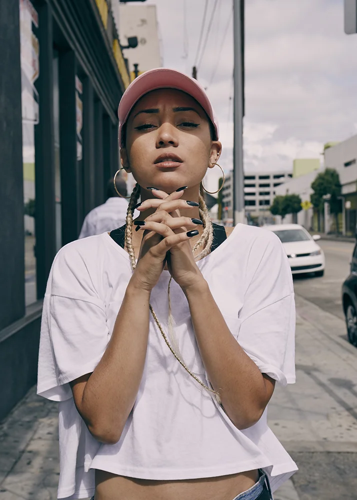 Young woman with braided hair, wearing a pink cap, large hoop earrings, and a white T-shirt, standing on urban sidewalk with buildings and cars in background.