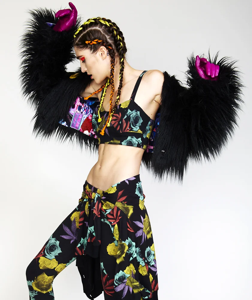 Young woman with braided hair in a colorful floral outfit, wearing a black furry jacket with pink gloves, posing with her head tilted and arms raised.