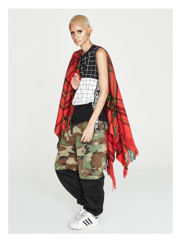 Fashion model wearing a red tartan blanket wrap, black and white grid-patterned sleeveless shirt, camouflage cargo pants, black joggers, and white Adidas sneakers against a plain white background.