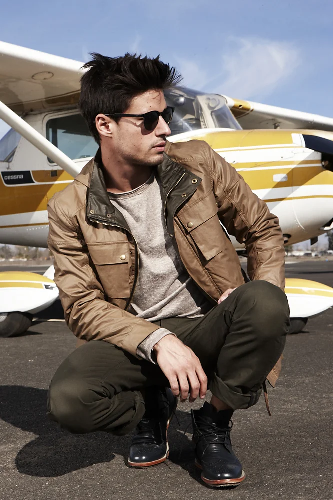 A young man squatting on an airport tarmac in front of a small yellow and white airplane, wearing sunglasses, a brown jacket, gray shirt, black pants, and black boots.