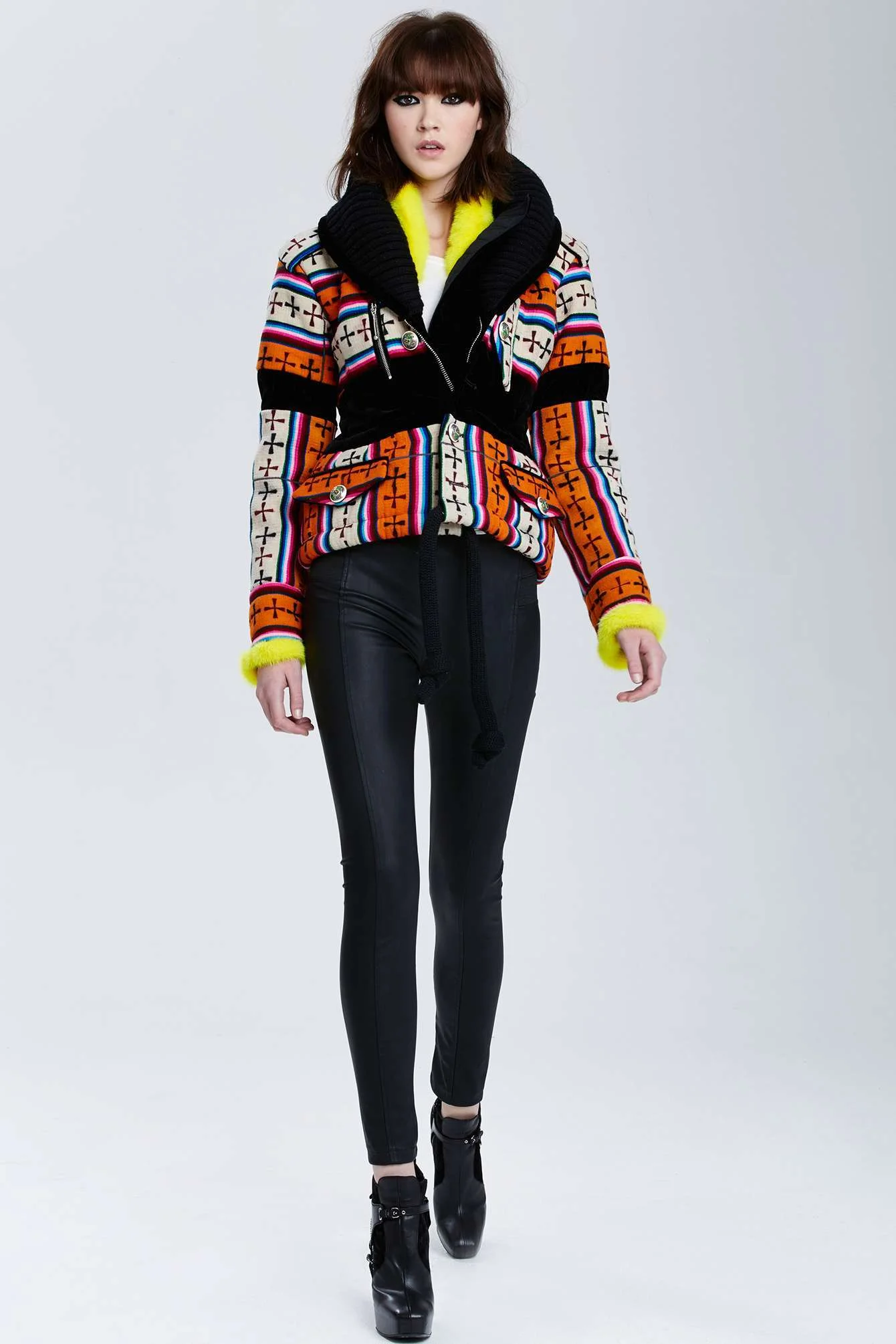 A woman wearing a colorful patterned jacket with yellow accents, black pants, and high-heeled shoes, standing against a plain background.