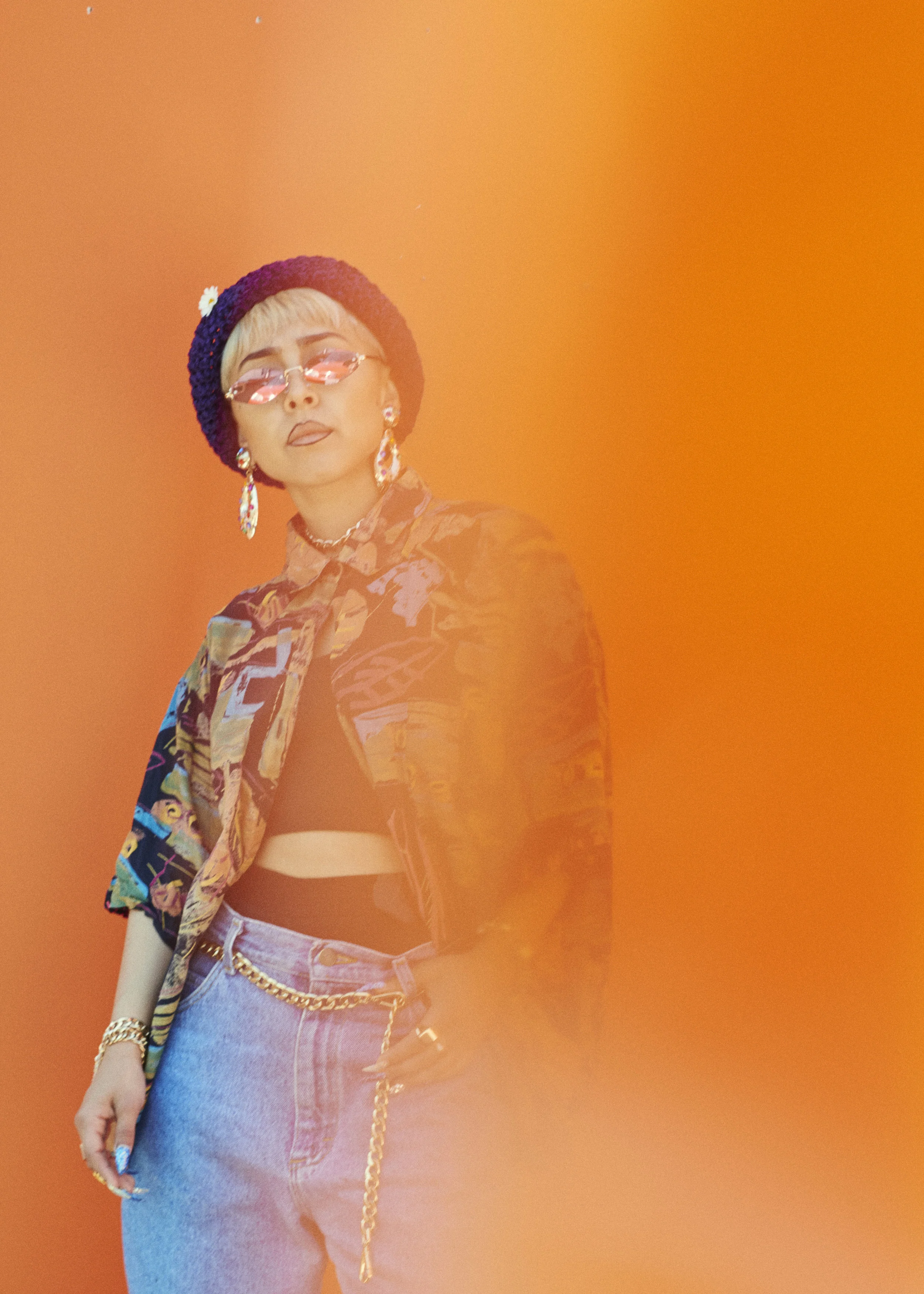 A woman wearing a colorful patterned jacket, light-wash jeans with a chain belt, a black crop top, a purple beanie with a flower, pink sunglasses, and large earrings, standing against an orange background with a blurred orange shape in the foreground