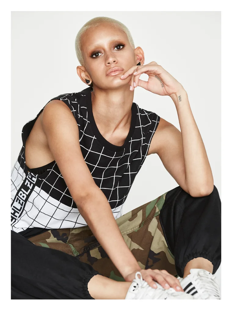 A young person with bleached blonde hair, wearing a sleeveless black and white grid-patterned shirt and camouflage pants, sitting on the floor with one hand resting on their knee and the other hand near their face, looking at the camera.