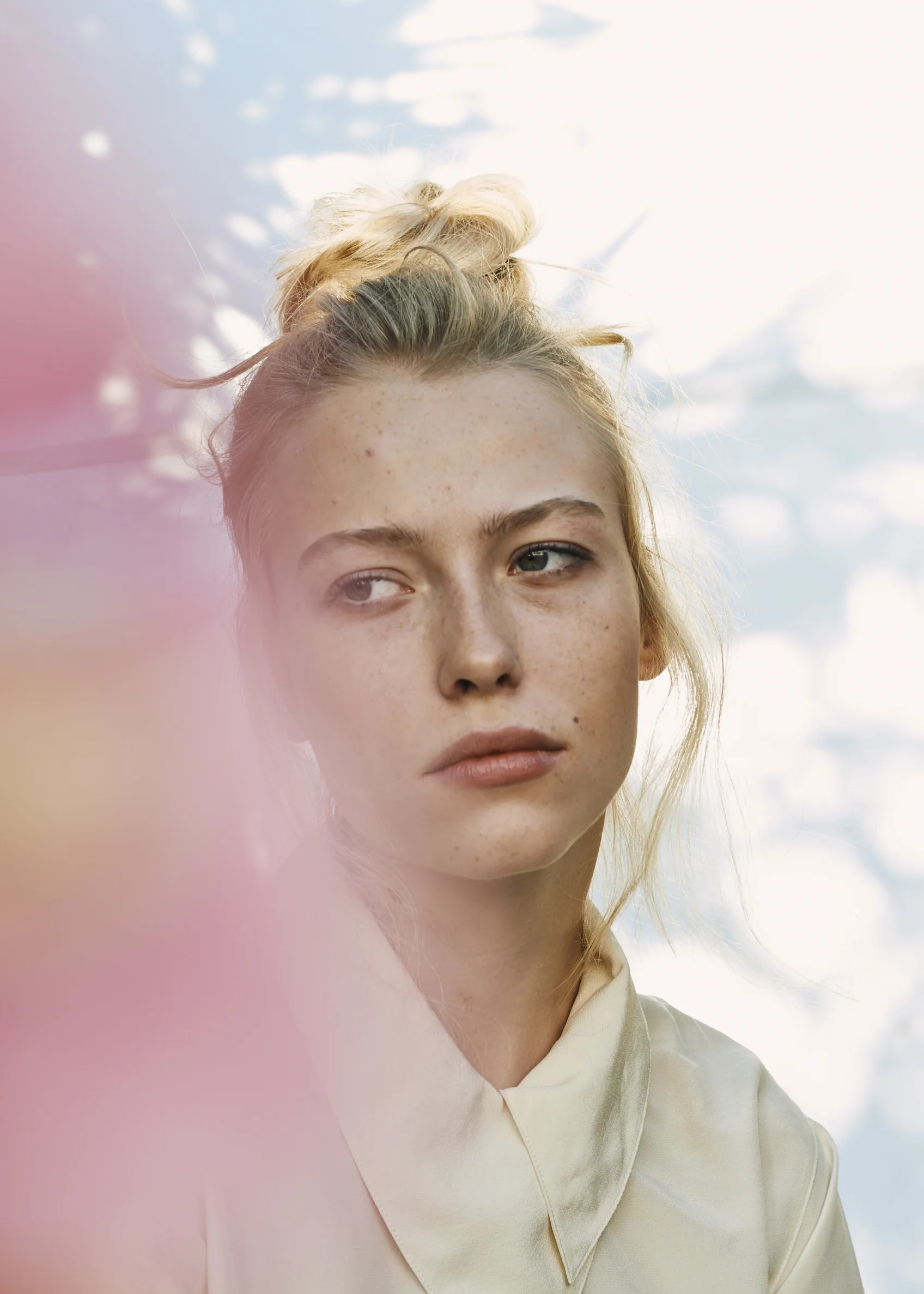 Close-up of a young woman with blonde hair styled in a messy bun, looking to the side with a neutral expression against a sky background with clouds.