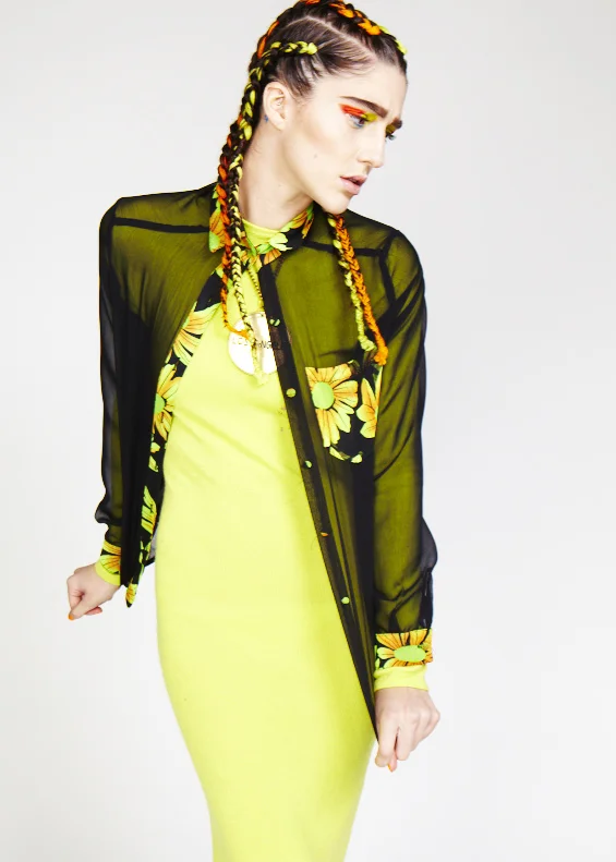 A woman with multicolored braided hair wearing a black sheer shirt with floral details over a lime green dress, standing against a plain background.
