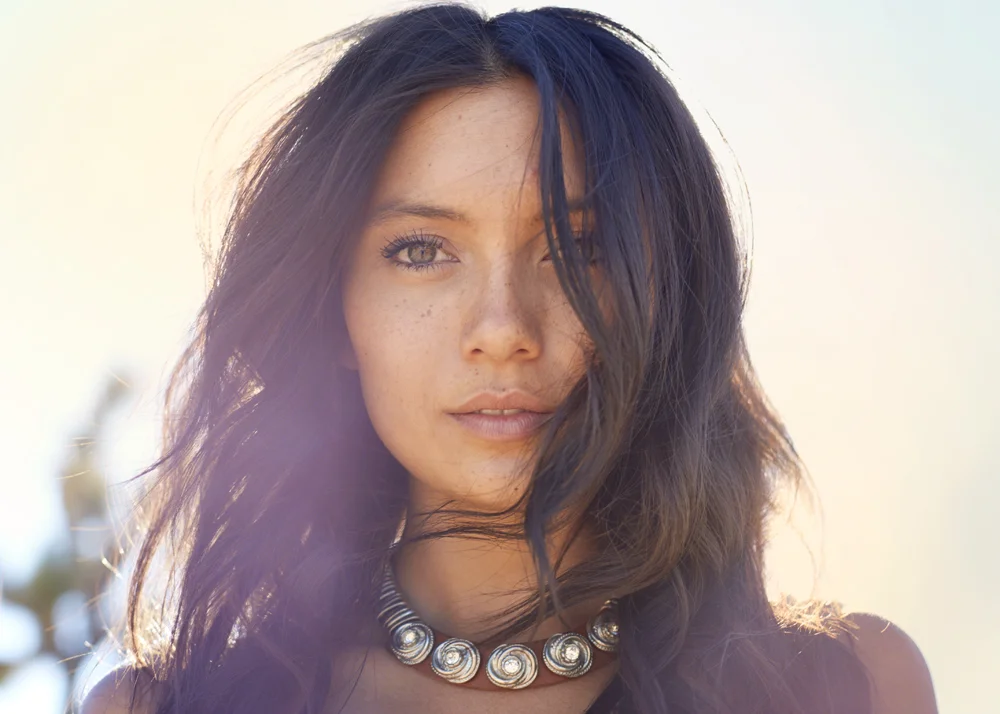 Close-up of a woman with long, wavy dark hair, wearing a silver shell necklace, outdoors with bright sunlight.