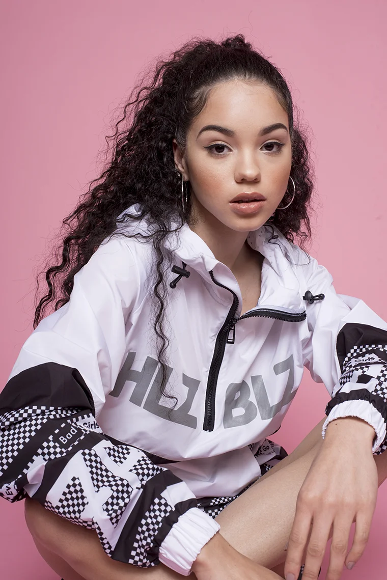 Young woman with long curly hair wearing a white and black track jacket, sitting against a pink background.