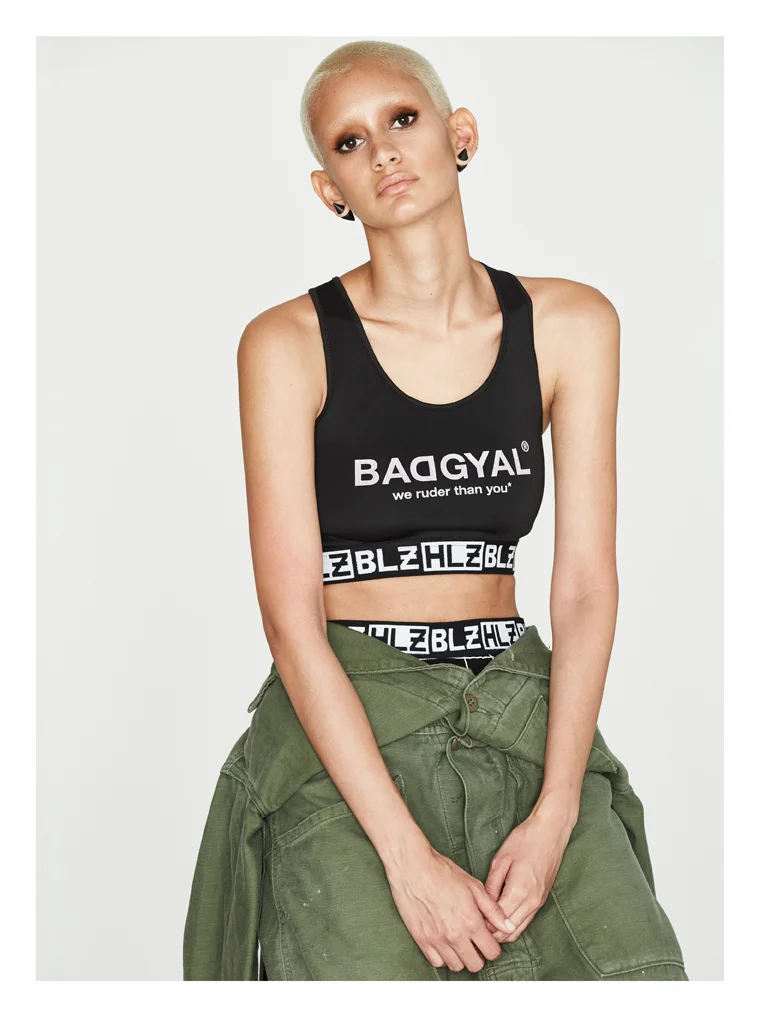 A young woman with short blonde hair, dark eye makeup, and multiple earrings, wearing a black crop top with white text and green pants around her waist.