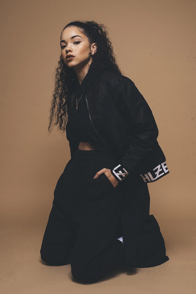 Young woman with curly hair and hoop earrings wearing a black bomber jacket and black pants, kneeling against a beige background.