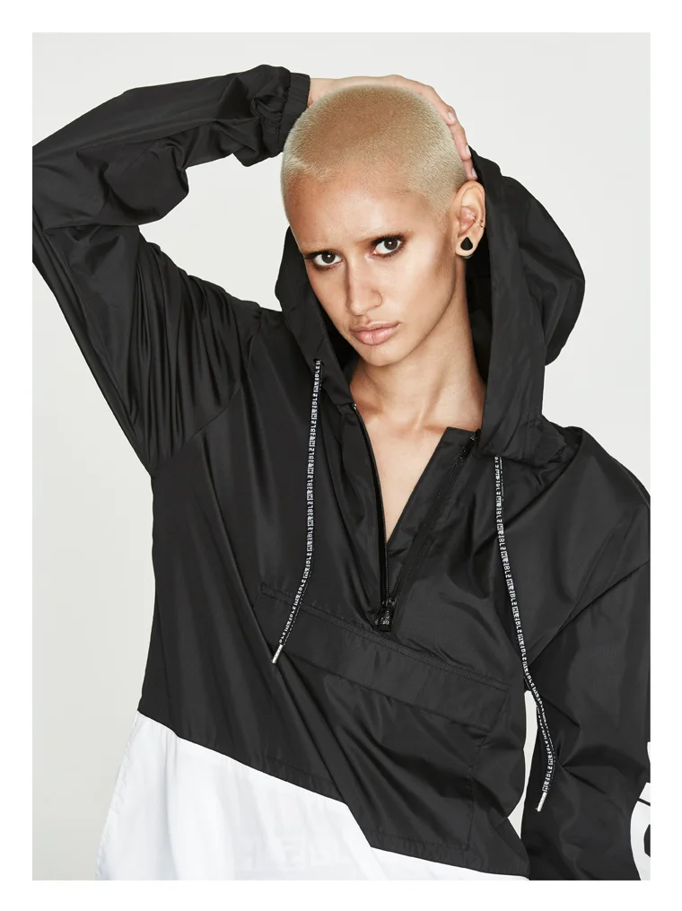 Woman with short blond hair wearing a black hooded windbreaker jacket and a white skirt, posing with one hand on her head against a plain light background.