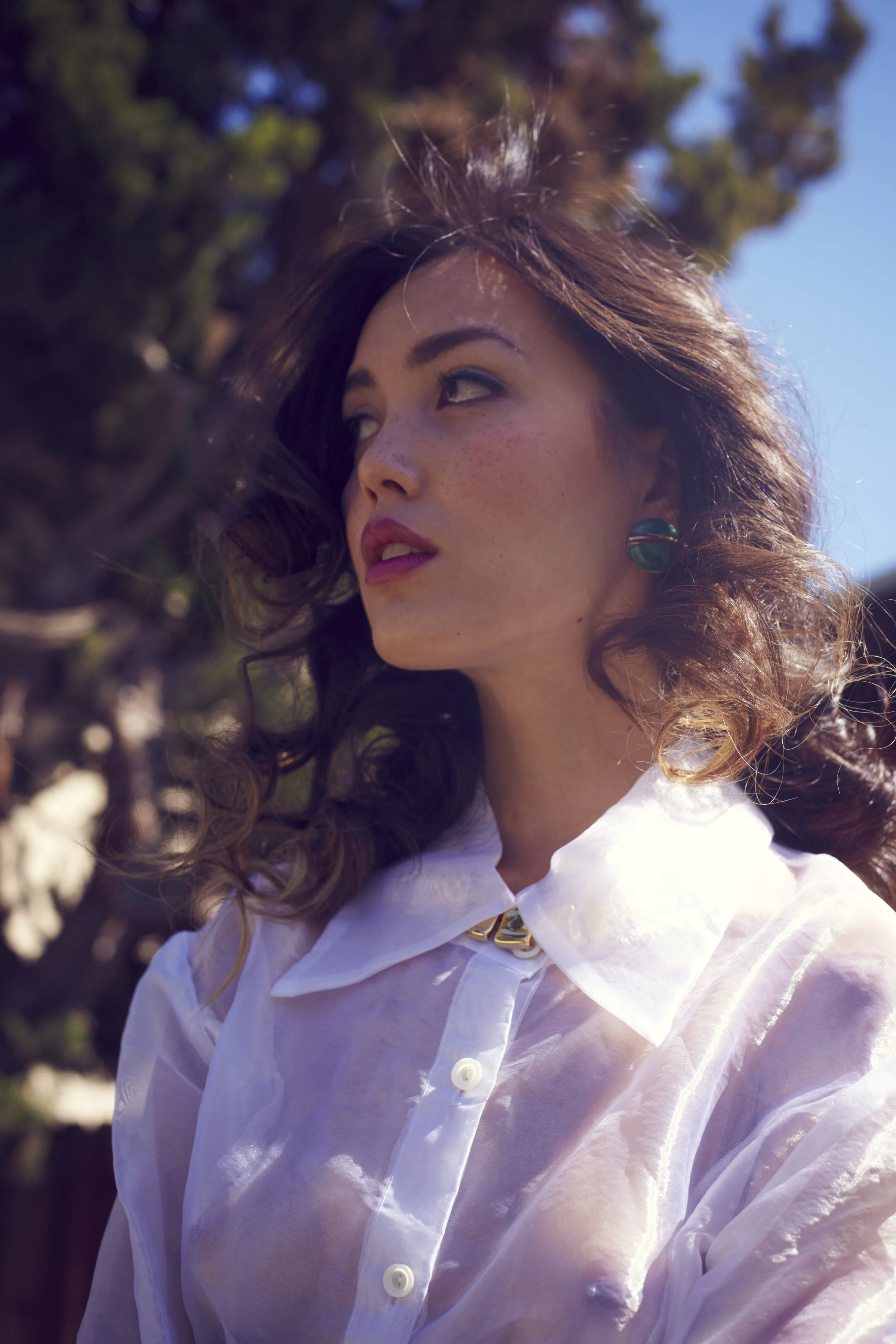 A woman with curly brown hair, wearing a white shirt and turquoise earrings, outdoors in sunlight with trees and a blue sky in the background.
