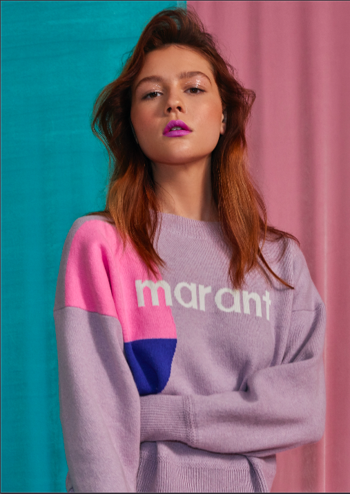 A young woman with wavy red hair and purple lipstick standing against a multicolored background with pink and teal curtains, wearing a lavender sweater with pink, purple, and blue color blocks, and the word "maraht" on it.