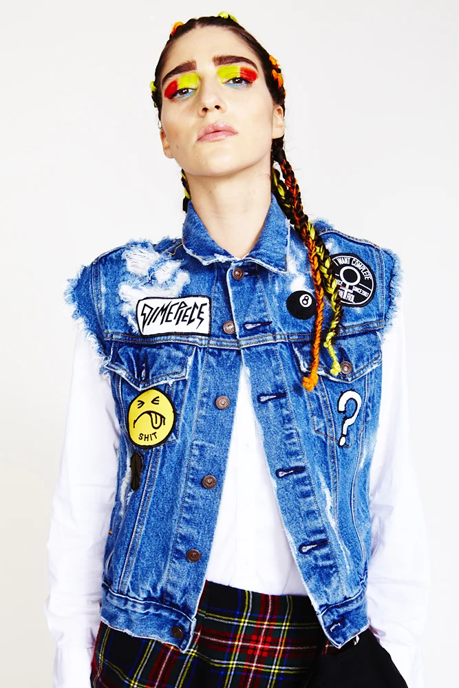 Woman with colorful eye makeup, wearing a denim jacket with patches, a white shirt, and plaid pants, against a plain white background.
