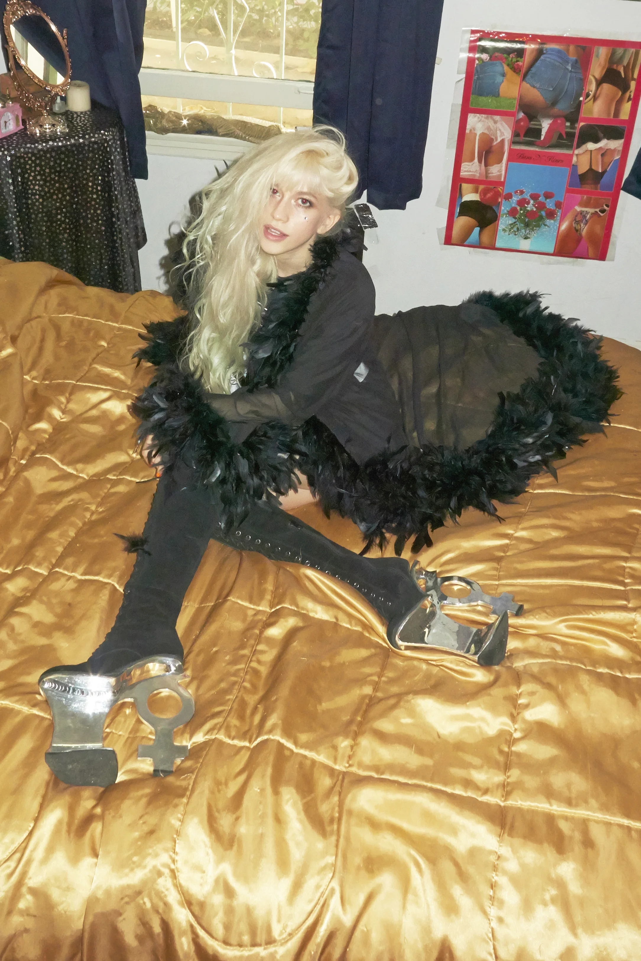 A woman with long blonde hair posing on a bed with a gold satin blanket, holding a large metallic sex toy shaped like a key with a female symbol. She is wearing black clothing with feathered details and black thigh-high boots.
