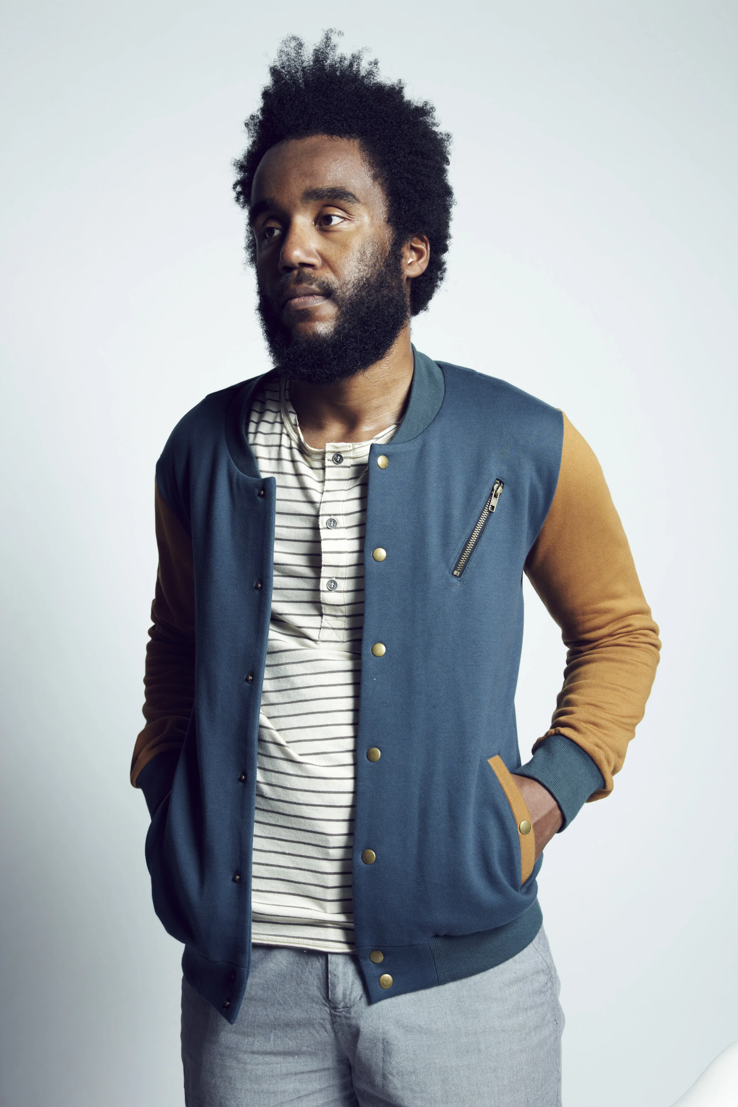 A man with a beard and curly hair wearing a navy and tan varsity jacket over a striped shirt, standing against a plain light background.