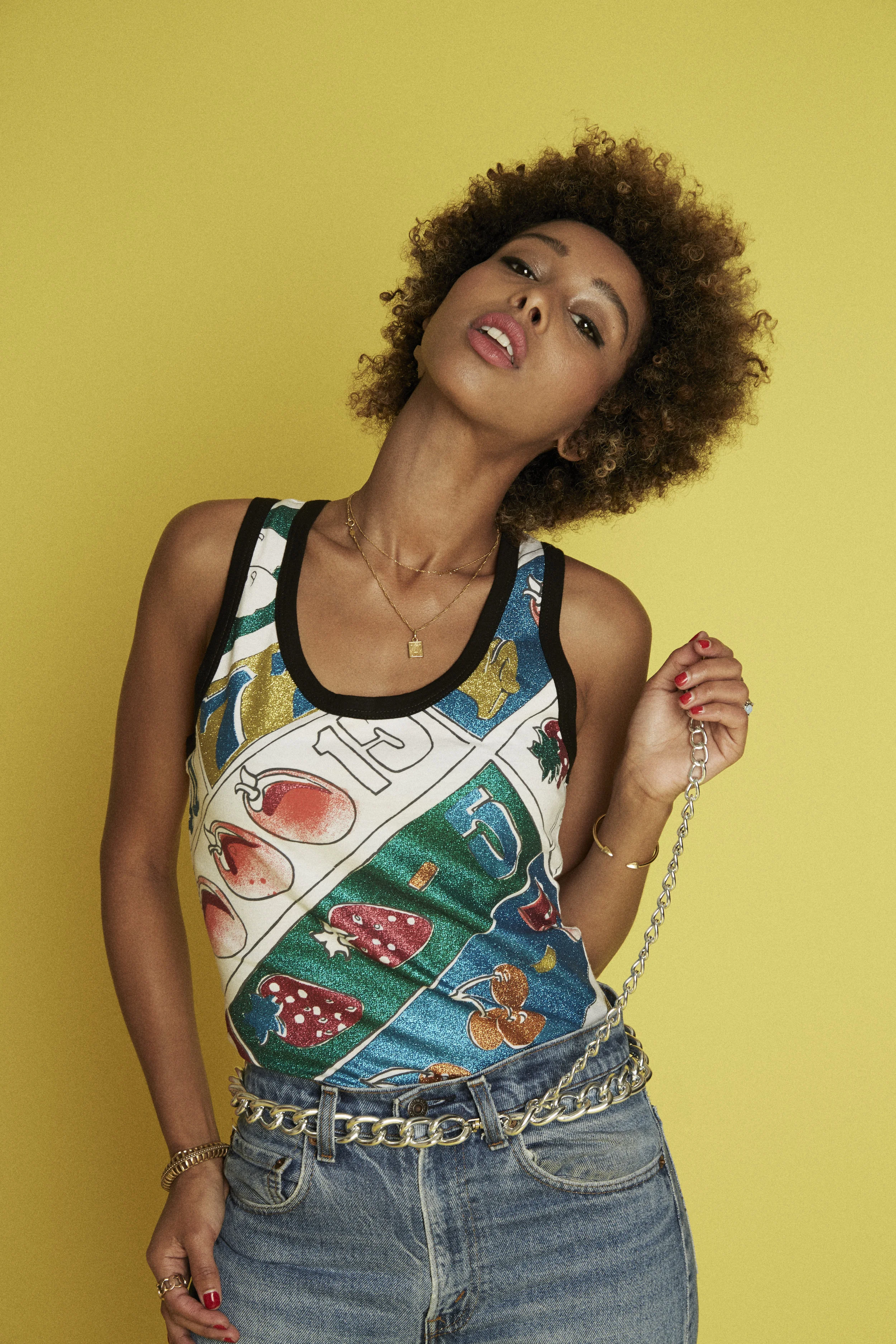 A woman with curly hair standing against a yellow background, wearing a colorful top with fruit designs, denim shorts, gold jewelry, and holding a chain purse.