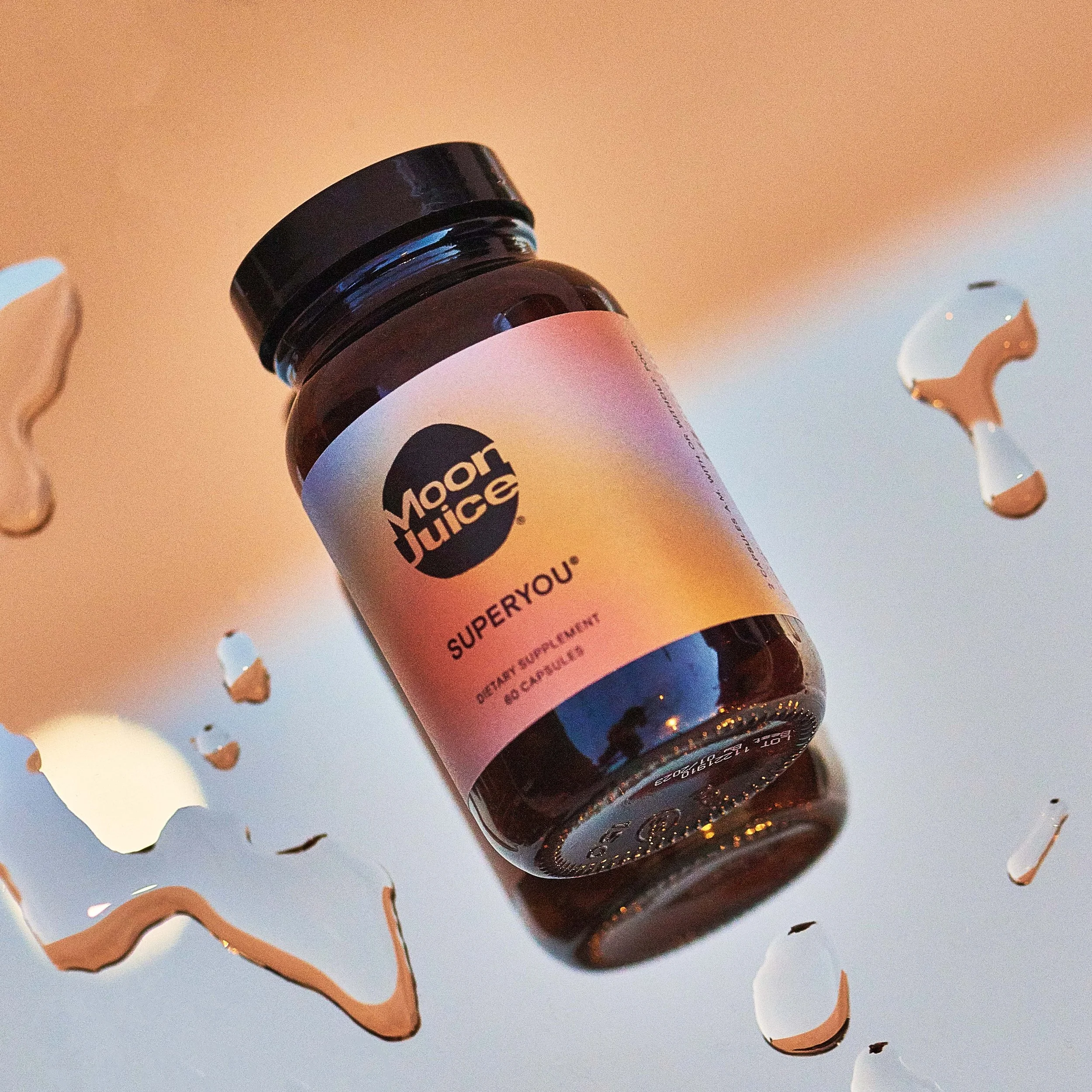 A bottle of Moon Juice SuperYou dietary supplement capsules surrounded by drops of liquid on a reflective surface with warm lighting.