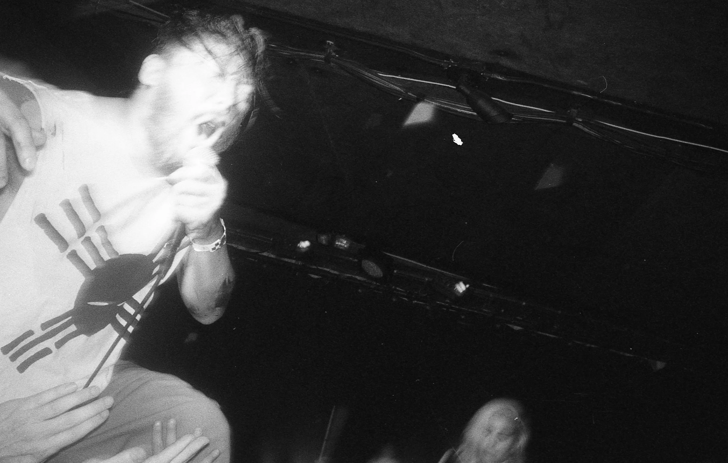 Person with long hair and tattoos on arms, wearing a t-shirt with a logo, holding a cigarette near their mouth, in a dark indoor setting with visible cables and wires on the wall.