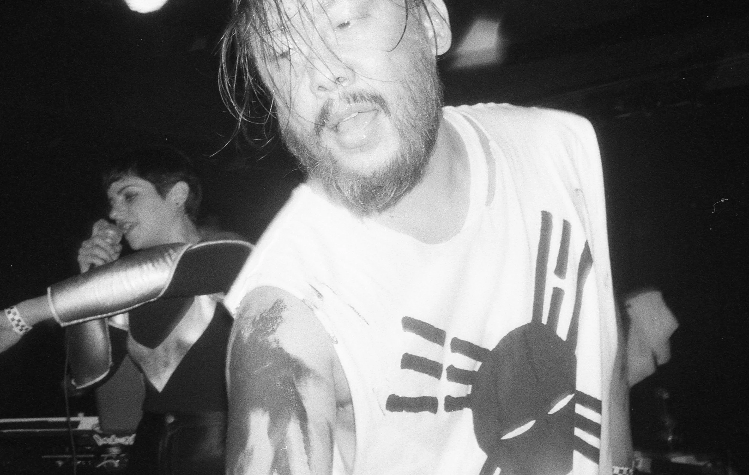 A black and white photo of a man with long hair, beard, and a sleeveless T-shirt with a graphic of a hand making a peace sign. He appears to be in a casual setting with another person in the background, who is smiling, drinking from a bottle, and has