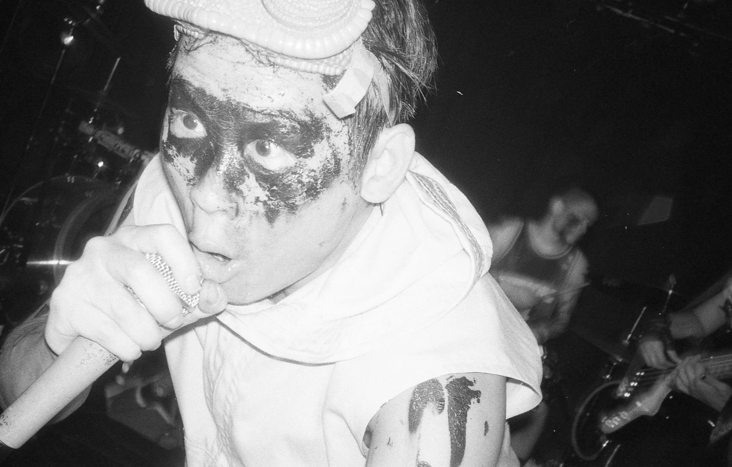 A person with face paint and a vape in their mouth, dressed in a light-colored top with a sleeve rolled up, is in a dark environment with others in the background.