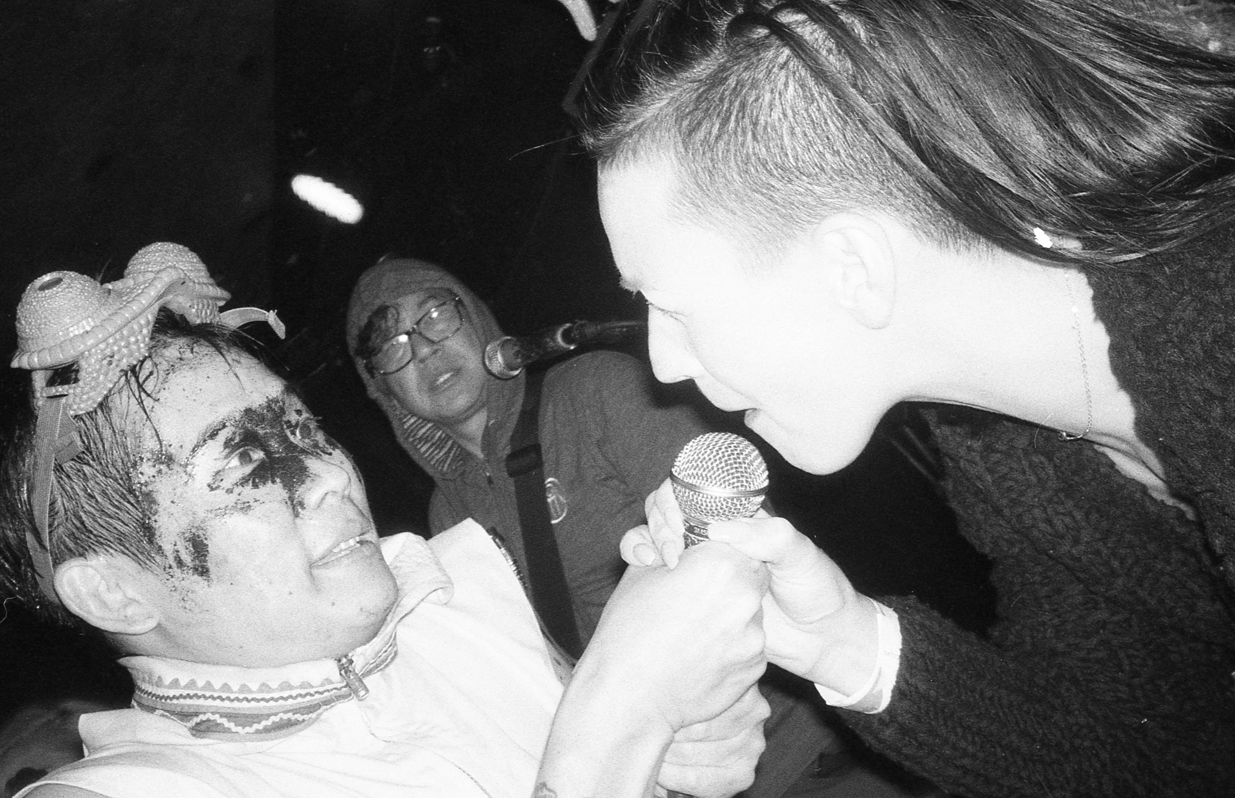 A woman dressed in clown makeup and a costume with a headpiece with tentacles is holding a microphone and looking at a person who is also holding a microphone, in an indoor setting with a dark background and other people watching.
