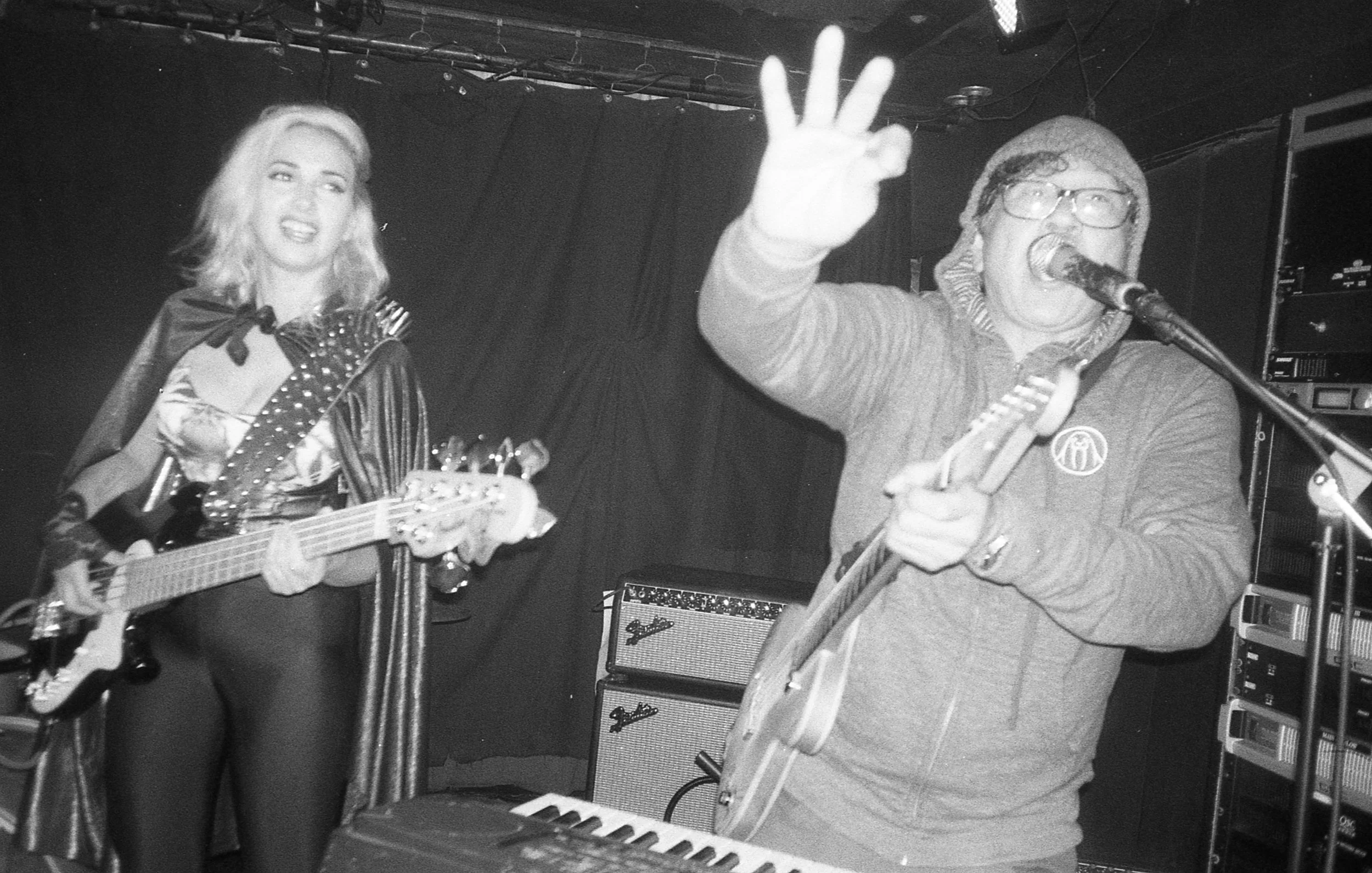 Black and white photo of a woman playing an electric guitar and a man singing into a microphone during a performance. The woman is smiling and wearing a cape, while the man is gesturing with his hand, wearing glasses, a beanie, and a hoodie. Musical 
