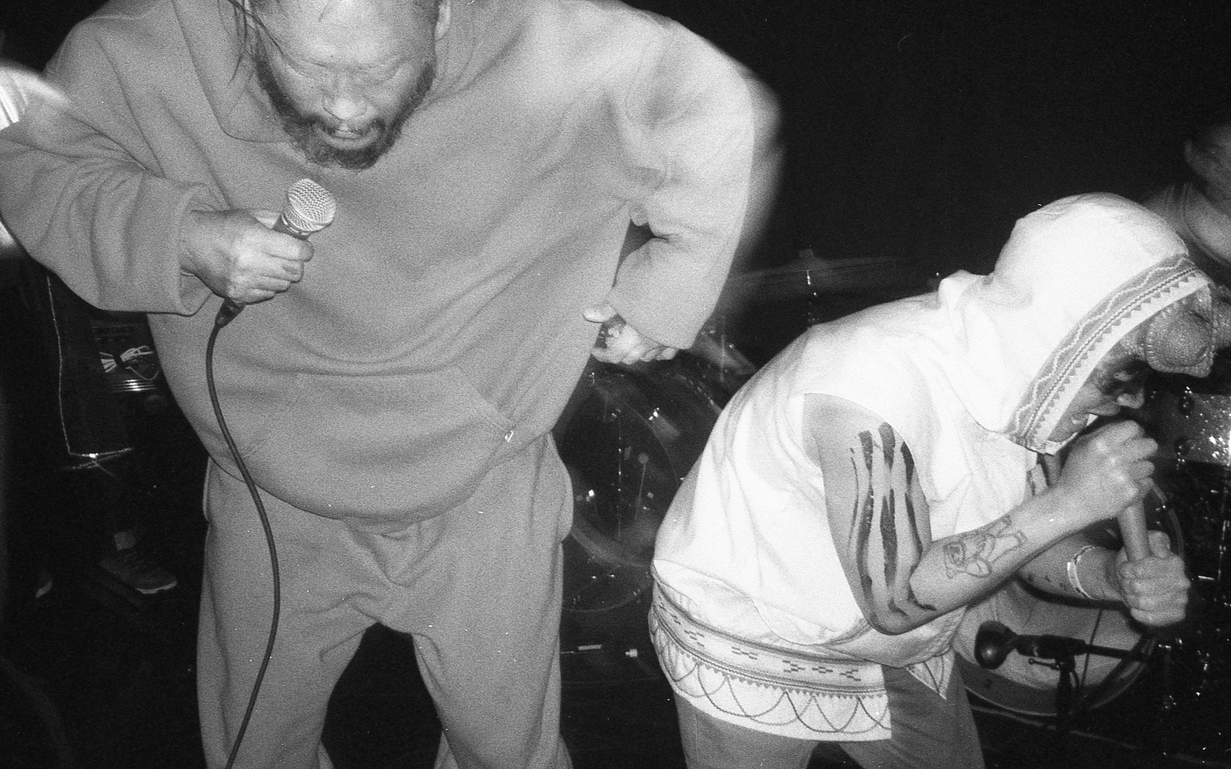 A man holding a microphone and singing or speaking passionately while another person, wearing a hoodie and sunglasses, leans into the microphone, in a lively scene likely at a music or entertainment event. music, editorial, album cover, photoshoot