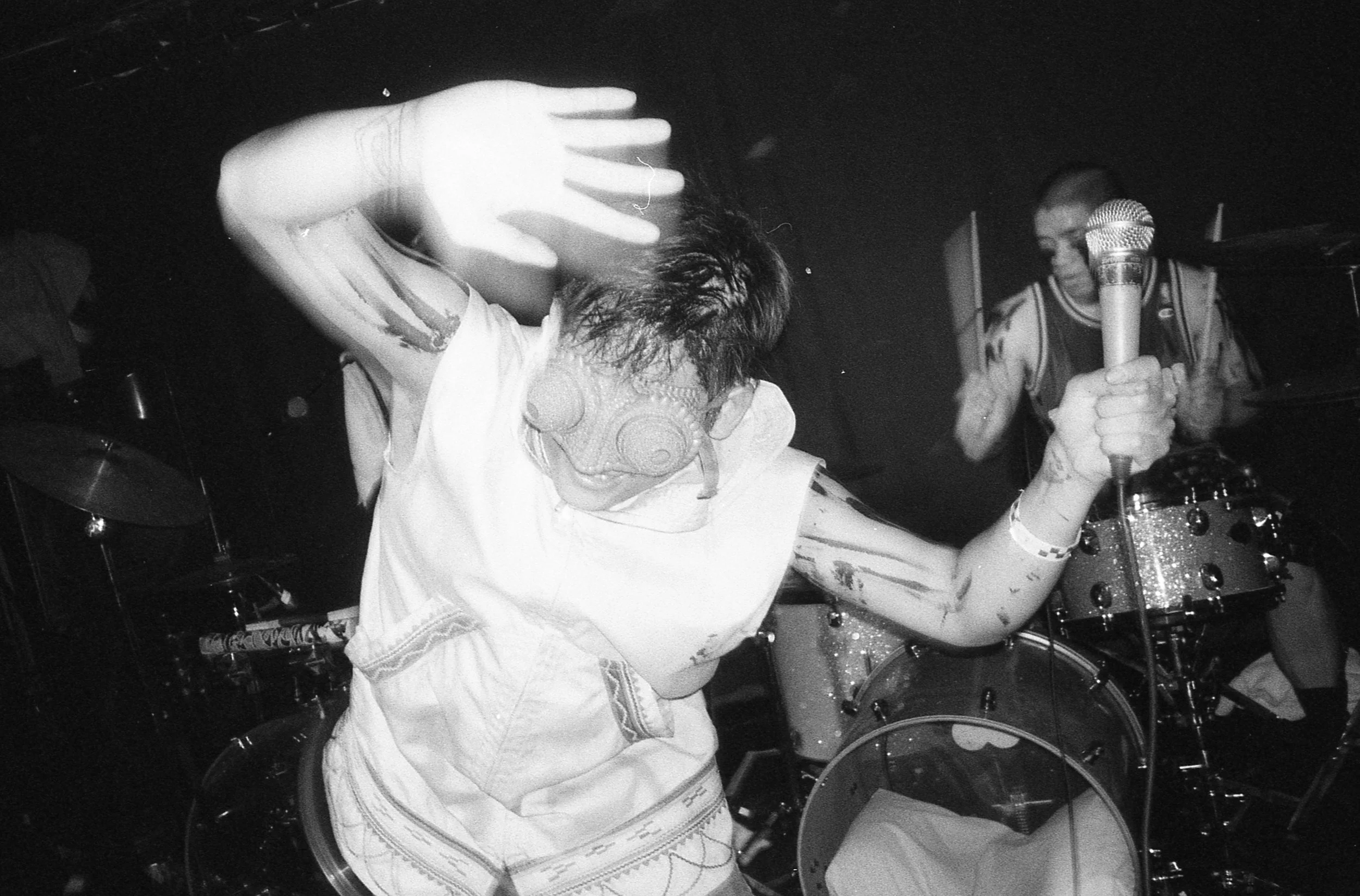 A person with tattoos dancing and holding a microphone on stage, with a drummer playing in the background. music, editorial, album cover, photoshoot, conceptual, concept art, David Choe