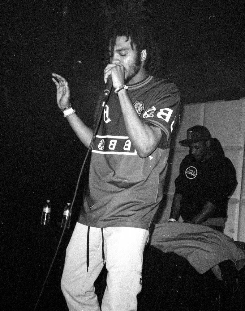 A man with dreadlocks singing into a microphone on stage, wearing a sports jersey and light-colored pants, with another person in the background wearing a cap and hoodie.