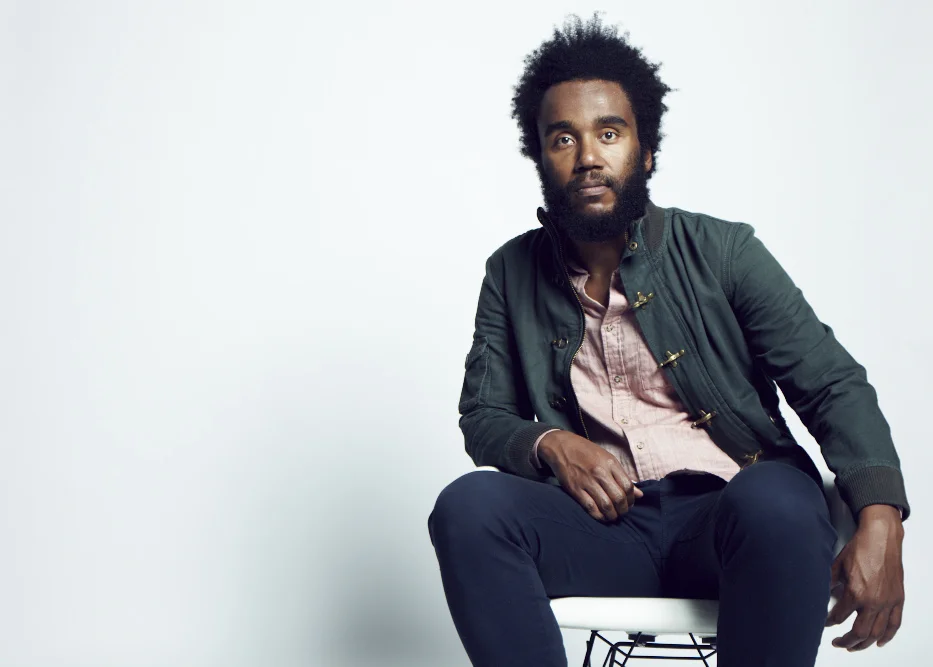 A man with curly black hair and a beard sitting on a white chair against a plain white background, wearing a dark green jacket and a pink shirt.