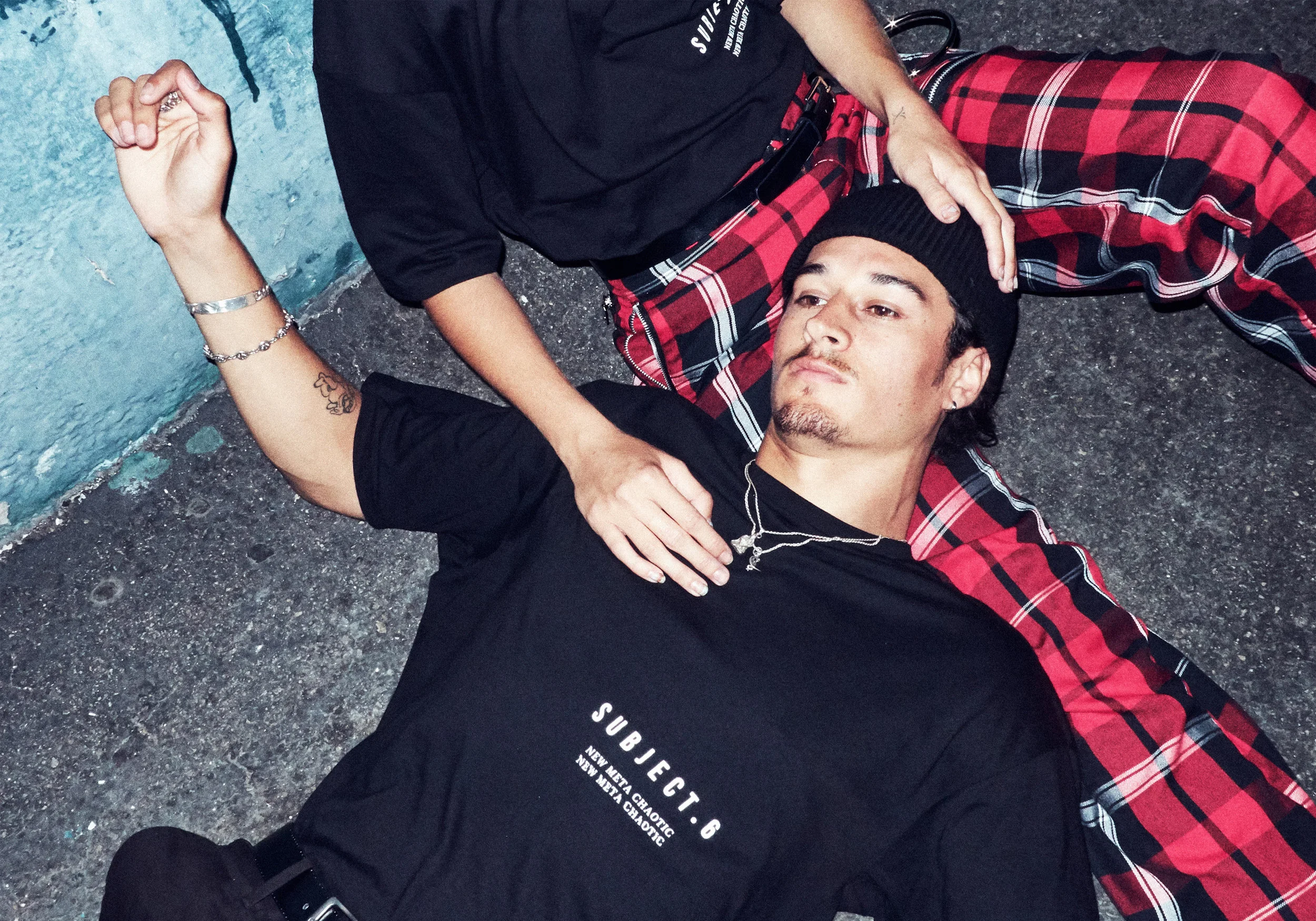 A young man lying on the ground against a blue wall, wearing a black beanie, black T-shirt with white text, and plaid pants. Another person is beside him, with one hand on his face and the other on his chest.