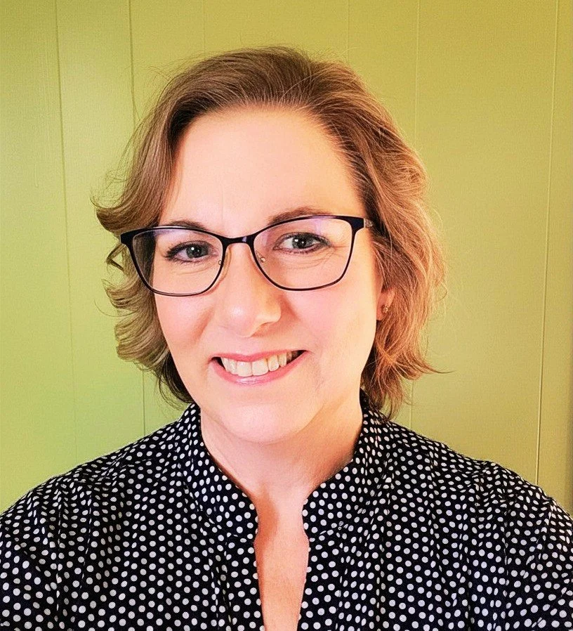 A woman with short, wavy brown hair, wearing glasses and a black and white polka dot blouse, smiling in front of a yellow wall.