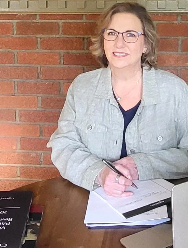 A woman with glasses sitting at a wooden table with papers and a pen, in front of a brick wall background.