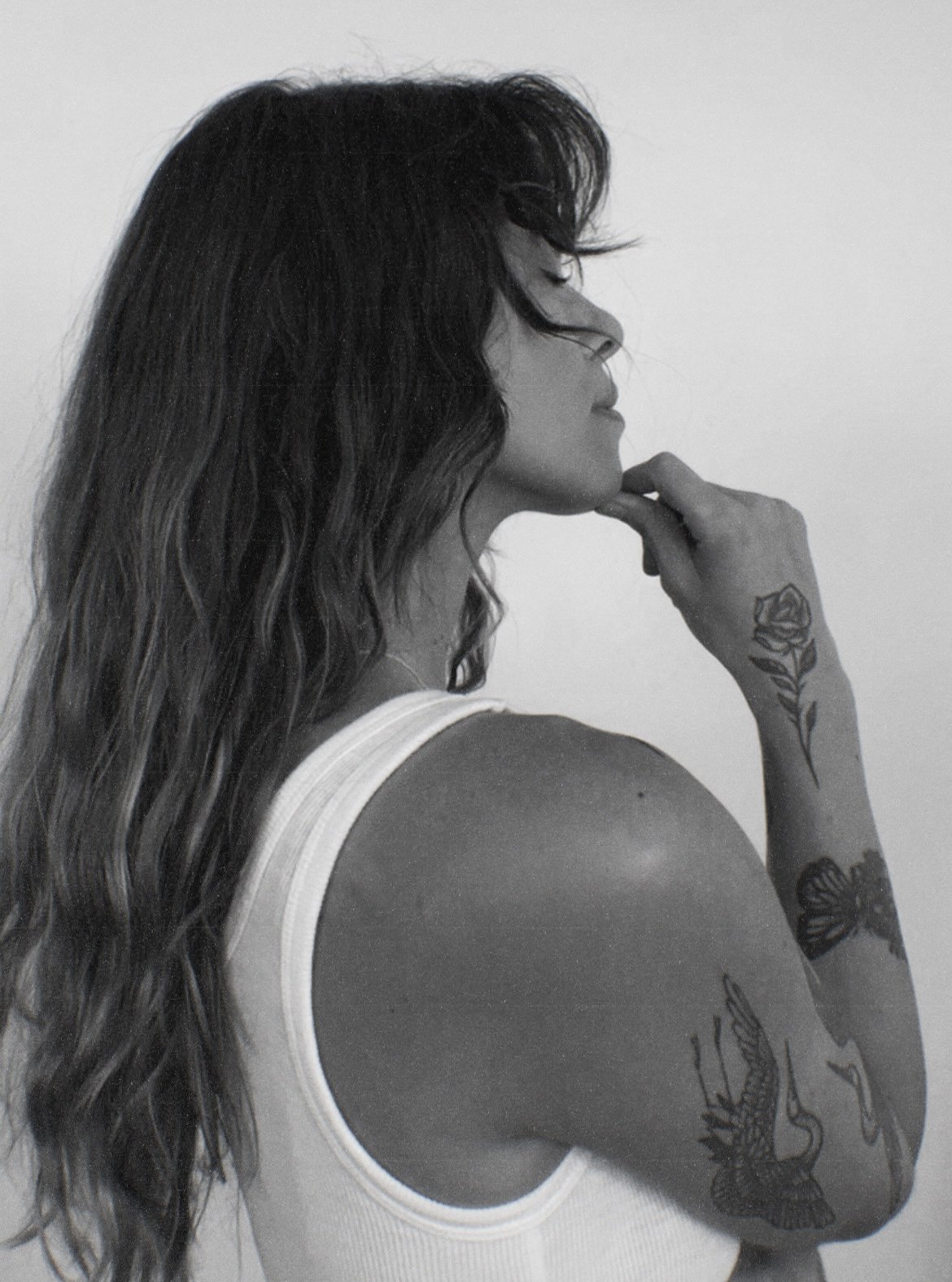 Black and white side profile of a woman with long wavy hair and tattoos resting her chin on her hand, POTS specialist supporting recovery from dysautonomia, MCAS, autoimmune, and post-viral illness.