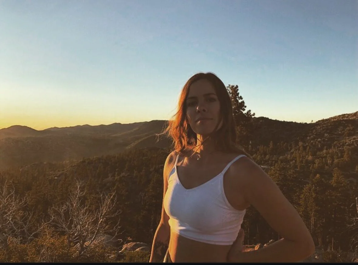 Woman with brown wavy hair wearing a white sports bra andstanding outdoors on a mountain overlook at sunset, POTS specialist supporting recovery from histamine intolerance, Lyme disease, autoimmune, and post-viral syndromes.
