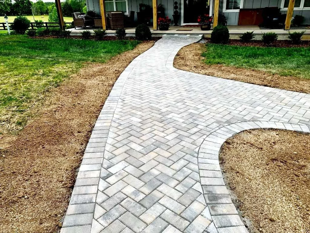Curved brick sidewalk leading to a house porch, with a landscape of green grass and shrubs on the sides.