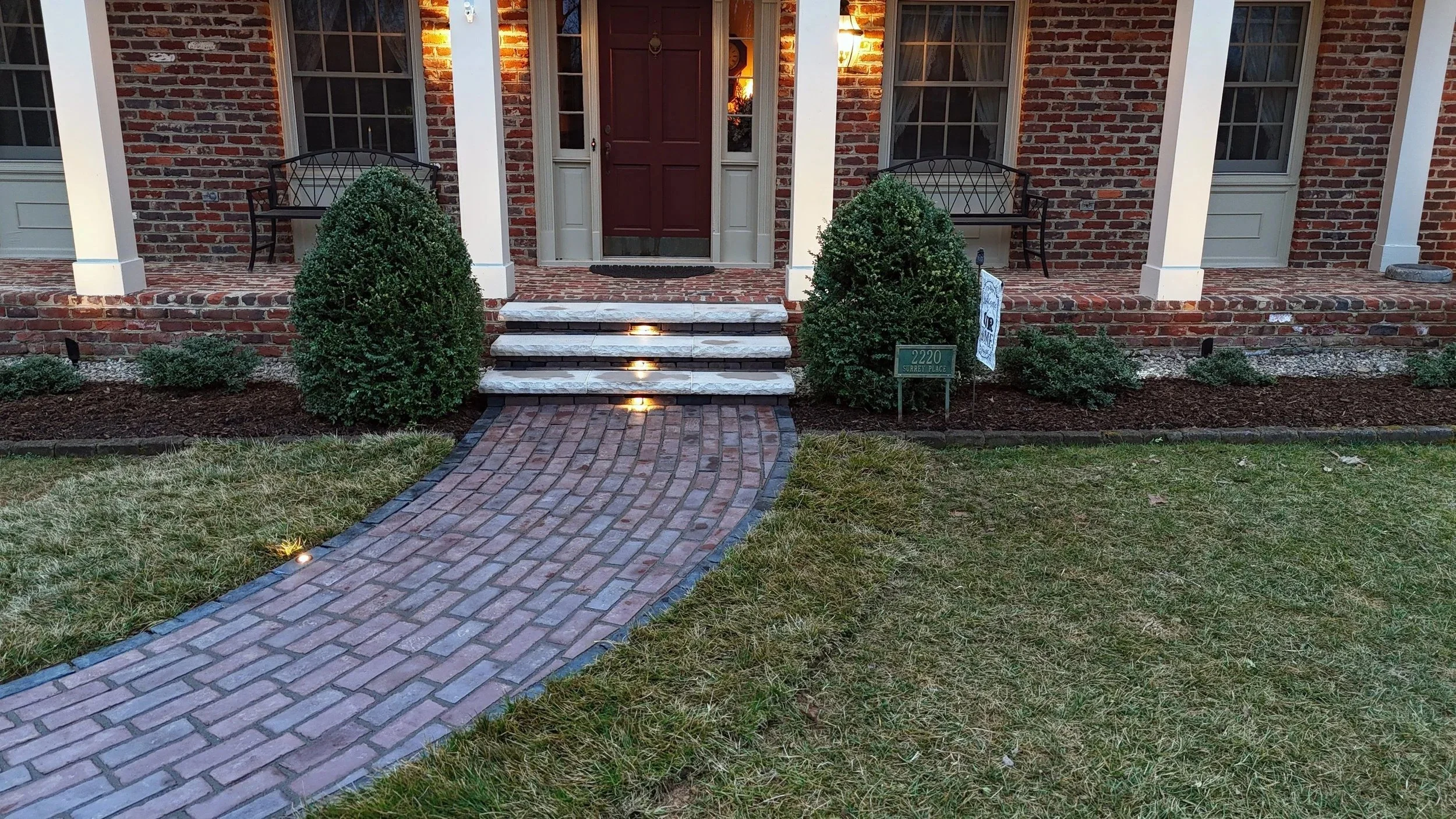Front porch of a brick house with a brick walkway, four steps with white stone treads, two bushes on each side, and illuminated pathway lights. The house has a red door, two black benches, and exterior lighting.