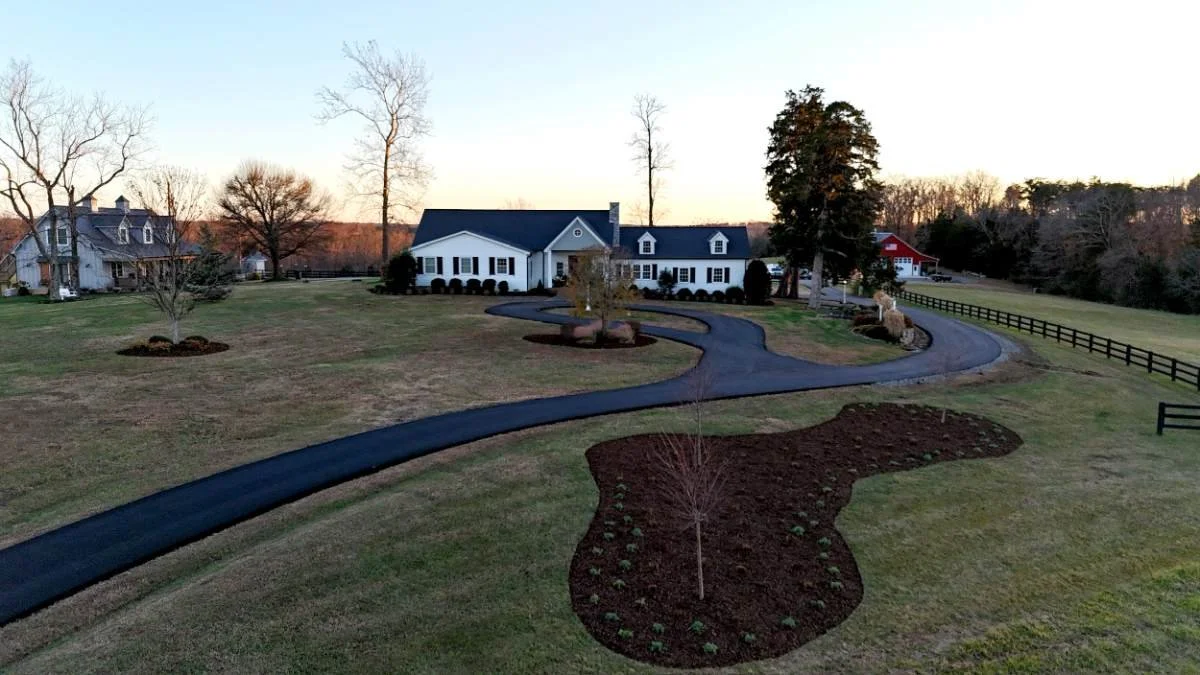 A winding paved driveway leading to a large white house with black shutters and a blue roof. The lawn has small trees and flower beds, with a black wooden fence on the right side. The sky is clear.