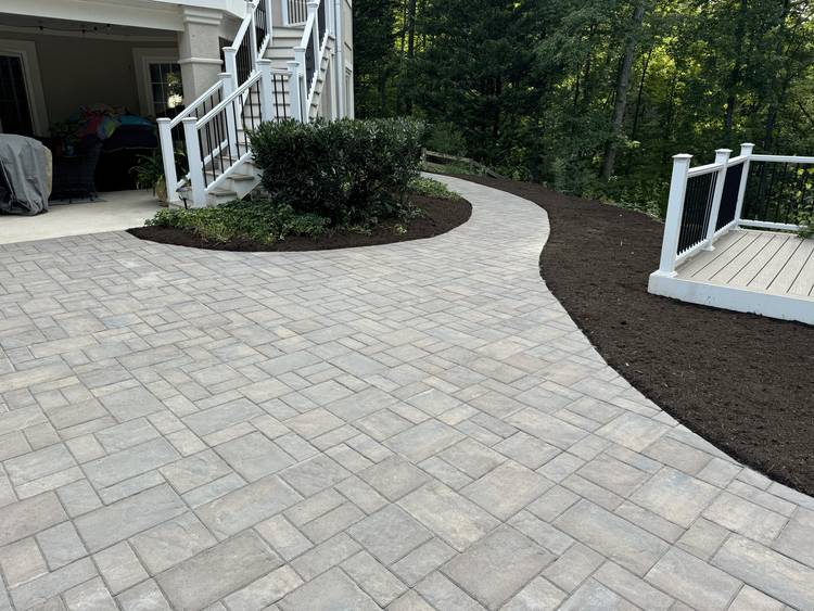 Newly paved stone patio with a small shrub in the center, bordered by dark mulch, near a white staircase and deck with a wooded yard background.