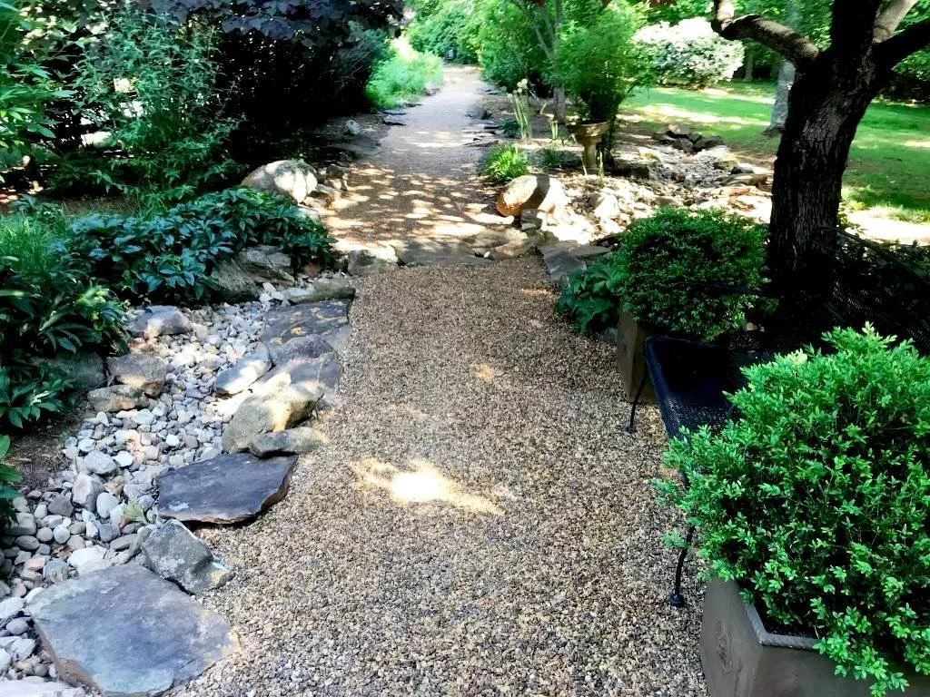A garden pathway with gravel, bordered by plants and trees, leading into a shaded lush garden.