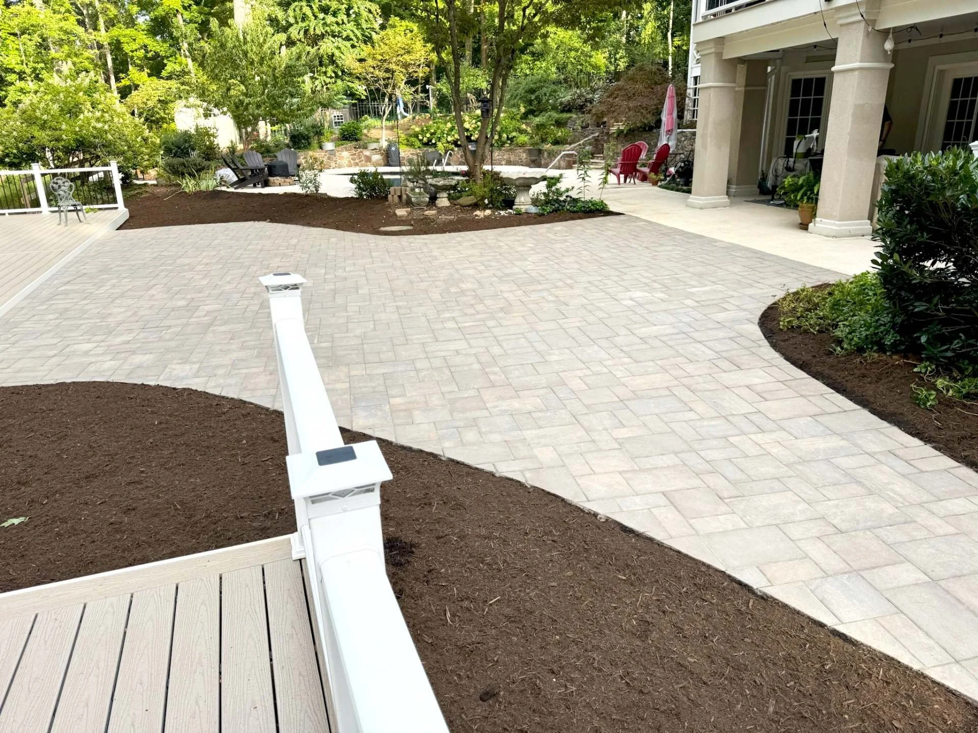 A backyard patio area with a brick paver surface, surrounded by landscaped flower beds and trees. There are outdoor chairs and a pool in the background, with a house on the right side featuring a covered porch.