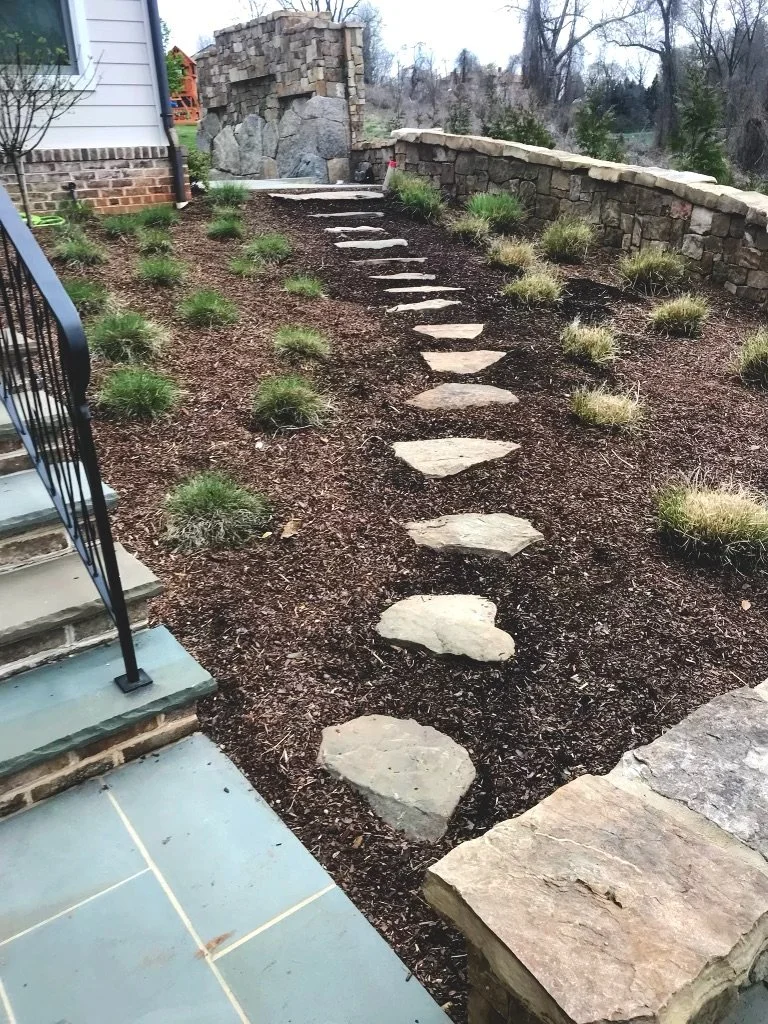 Stone pathway with stepping stones leading up a landscaped yard adjacent to a house with brick and siding exterior, and a stone retaining wall in the background.