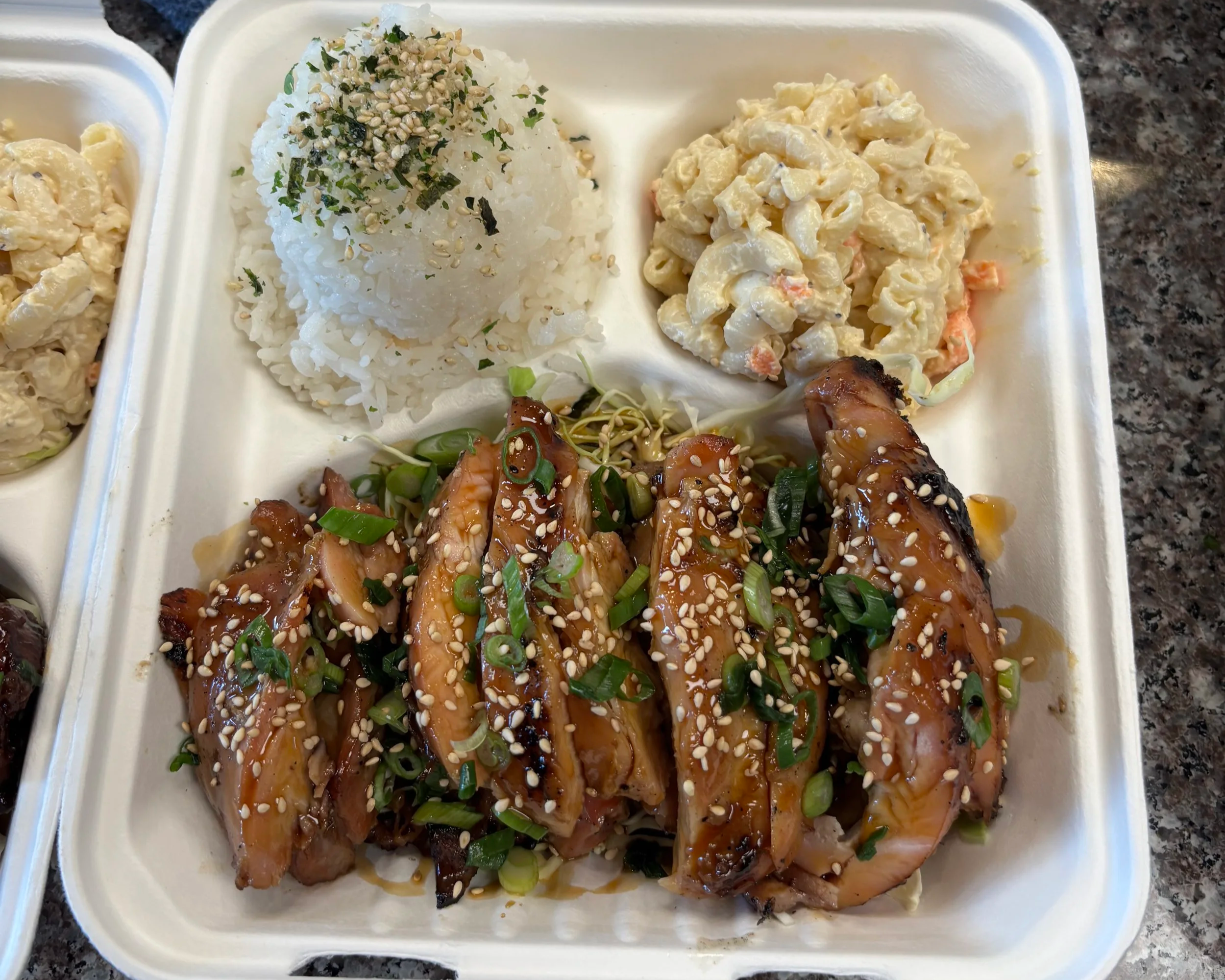 A divided takeout container with grilled chicken glazed with a teriyaki, sprinkled with sesame seeds and chopped green onions, served with a mound of rice topped with furikake, and hawaiian mac salad