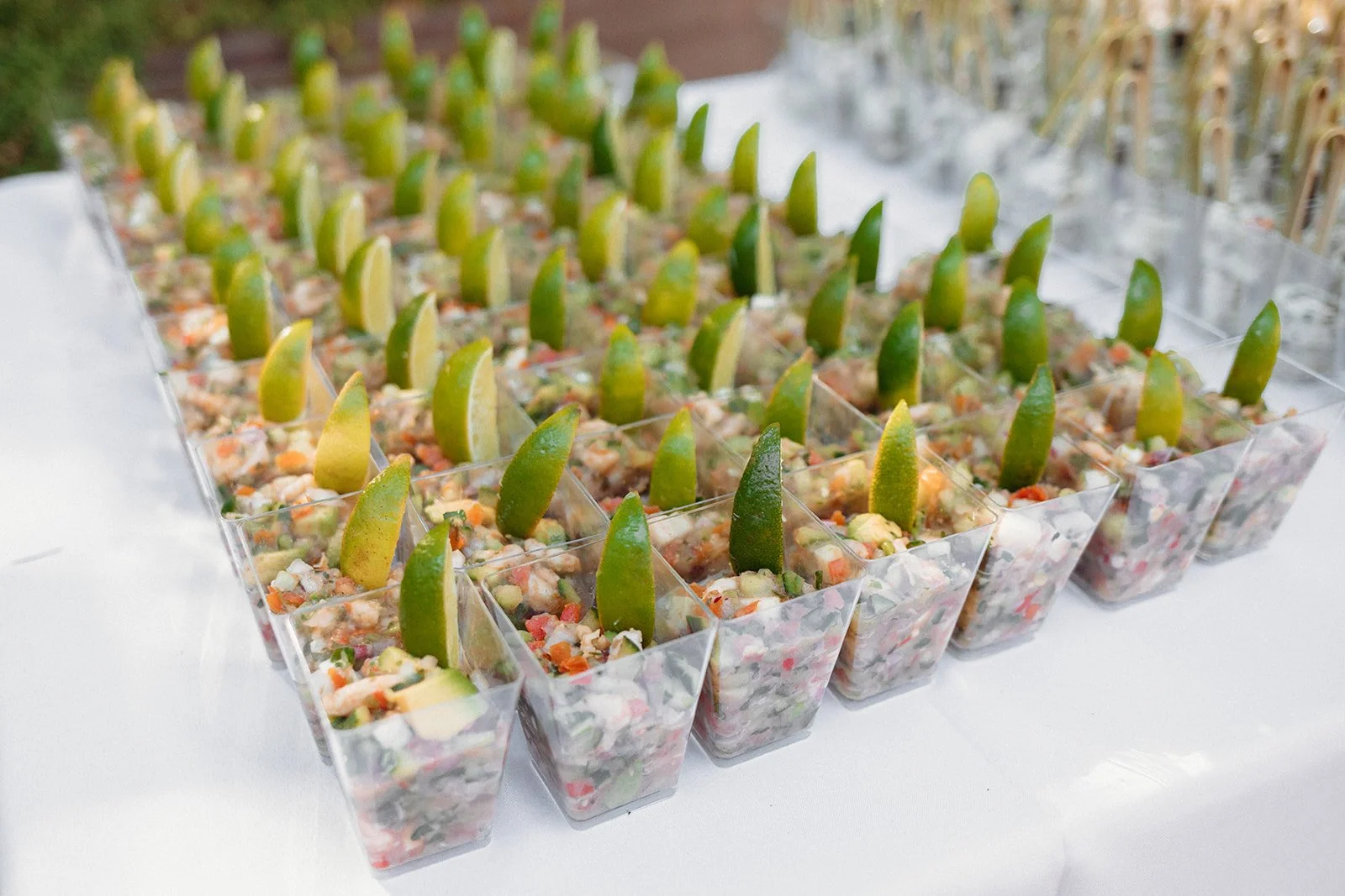 A large display of small clear cups containing a colorful salmon and avocado ceviche topped with a lime wedge.